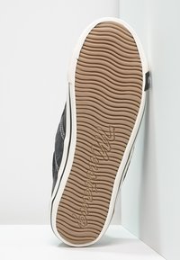 Sole černého tenisky s bílým lemem, ukazující vlnitý vzor podrážky a vytlačené jméno značky, umístěná vertikálně proti bílému rohu.