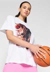 Bílý tričko s grafickým potiskem basketbalových bot, nošené s růžovými šortkami, držící oranžový basketbal. Měkký bavlněný materiál a uvolněný střih.
