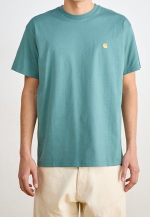 Hombre con una camiseta lisa de cuello redondo color azul verdoso con un pequeño logo amarillo en el pecho izquierdo y pantalones de color crema.
