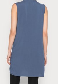 Tunic top blu senza maniche in un tessuto morbido, con colletto, taglio dritto e orlo arrotondato, indossato su leggings neri.
