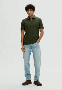 Νέος άνδρας στέκεται φορώντας ένα σκούρο πράσινο κοντομάνικο πουκάμισο πόλο, ανοιχτό μπλε τζιν και καφέ μπότες, με το ένα χέρι στην τσέπη.