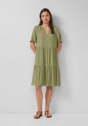 Junge Frau trägt knielanges olivgrünes Stufenkleid und braune Sandalen und steht vor einer schlichten hellgrauen Wand.