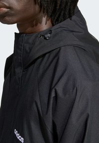 Svart vattentät jacka med hög krage, utrustad med dragskojustering och smal passform. Adidas-logga synlig framtill. Slät textur.