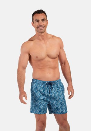 Uśmiechnięty muskularny mężczyzna stojący, ubrany w niebieskie kąpielówki z wzorem i czarnym sznurkiem, na jednolitym jasnym tle.