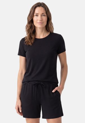 Vrouw met golvend bruin haar die een zwarte T-shirt met korte mouwen en zwarte short met trekkoord draagt, staand met één hand in de zak.