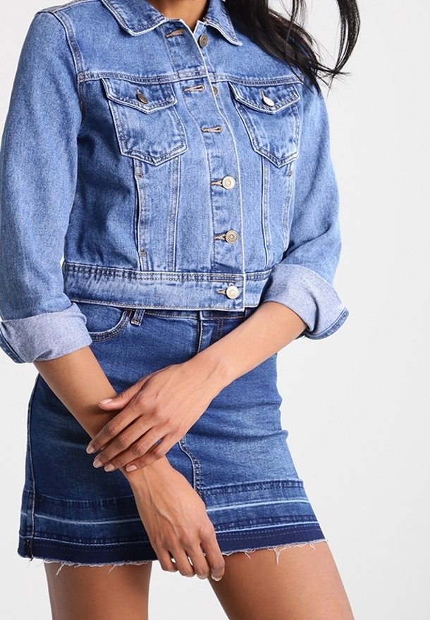 Jeansowa kurtka o wyblakłym, jasnoniebieskim wykończeniu, z czterema kieszeniami z przodu i guzikami. Nosi się ją na dopasowanej, niebieskiej jeansowej mini spódnicy z postrzępionym dołem.