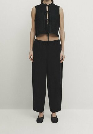 Black cropped top with knit texture and front tie detail, paired with loose-fitting black trousers and flat black shoes.