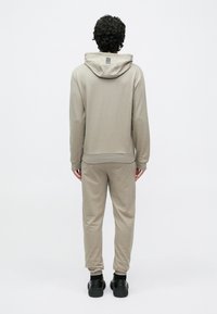 Személy bézs kapucnis pulóvert és hozzá illő melegítőnadrágot viselve, háttal áll egy egyszínű fehér háttér előtt.