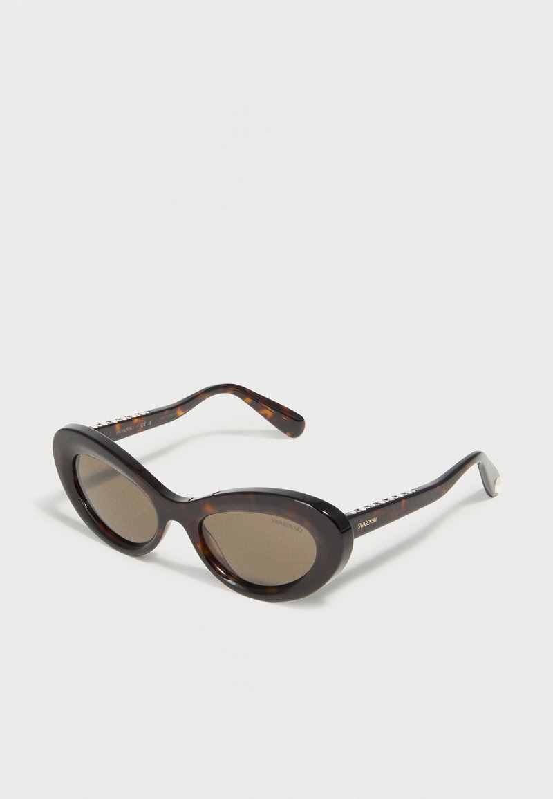 Γυναικεία γυαλιά ηλίου με σκελετό σε σχήμα γατίσιου ματιού και χρυσόχρωμο σχέδιο, με σκούρους φακούς και σήμανση Swarovski στα πλαϊνά, εκτεθειμένα σε λευκή επιφάνεια.