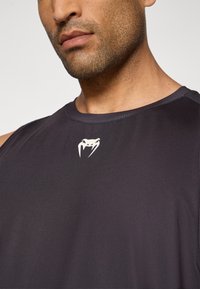 T-shirt de sport sans manches de couleur sombre, fabriqué en tissu lisse, avec un logo blanc ressemblant à la tête d'un serpent sur la poitrine.