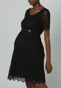 Czarna koronkowa sukienka z krótkimi rękawami, misternymi wzorami kwiatowymi, wyraźnie zaznaczoną talią z smukłym czarnym paskiem oraz falbaniastym wykończeniem dołu.