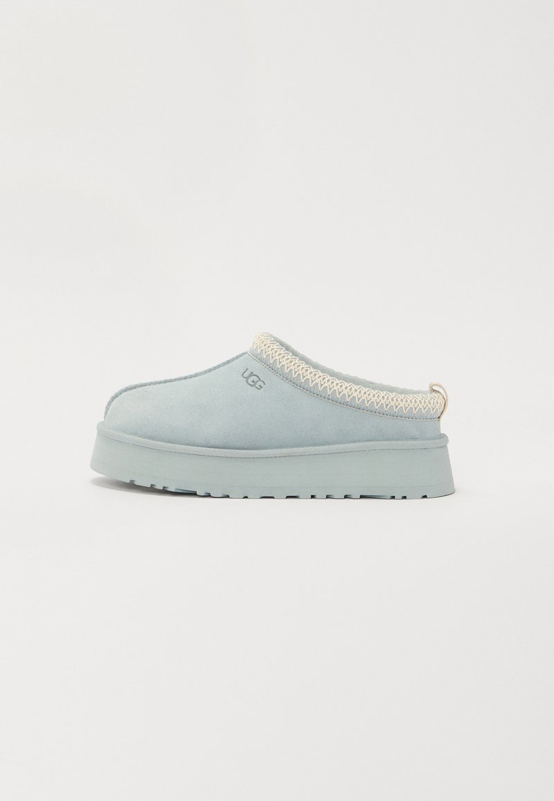 Lichtblauwe instapslippers met een dikke zool, witte gestikte rand en UGG-logo aan de zijkant, weergegeven op een effen witte achtergrond.