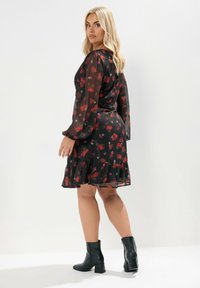 Robe fleurie avec de longues manches transparentes, base noire, motifs rouges et roses, ourlet en volants superposés. Accessoirisée d'une ceinture noire et de bottines.
