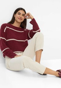 Kvinde sidder på gulvet iført en bordeaux sweater med beige striber, beige bukser og bordeaux sko, smilende og hviler sit hoved på sin hånd.