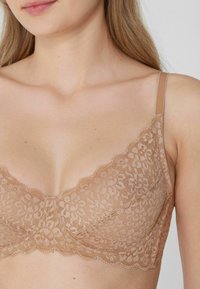 Núdový krajkový podprsenka s vlnitými okraji, květinovým vzorem, průhlednou tkaninou a nastavitelnými ramínky. Design má hladký, zakřivený tvar.