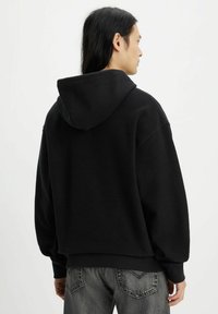 Hoodie oversize nero con una texture morbida, grande cappuccio e polsini a costine. Indossato con jeans scuri caratterizzati da un motivo sottile.