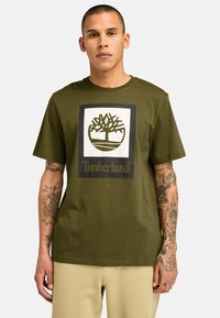 Olivově zelené bavlněné tričko s centrálním grafickým motivem loga stromu uvnitř čtverce a slovy "Timberland" v tučném písmu pod ním.