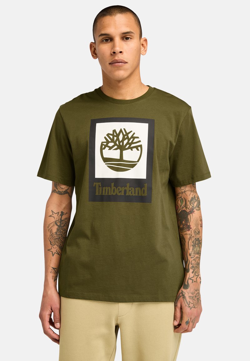 Olivově zelené bavlněné tričko s centrálním grafickým motivem loga stromu uvnitř čtverce a slovy "Timberland" v tučném písmu pod ním.