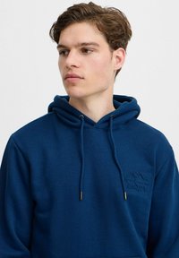 Navy blue hoodie in soft fabric features a kangaroo pocket, adjustable drawstrings with gold accents, and an embossed "Enjoy" logo.
