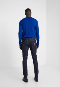 Modrý svetr, tmavé džínové kalhoty s logovou nášivkou, úzký střih, texturovaná látka, jednobarevné, nošeno s černými botami. Pohled zezadu.