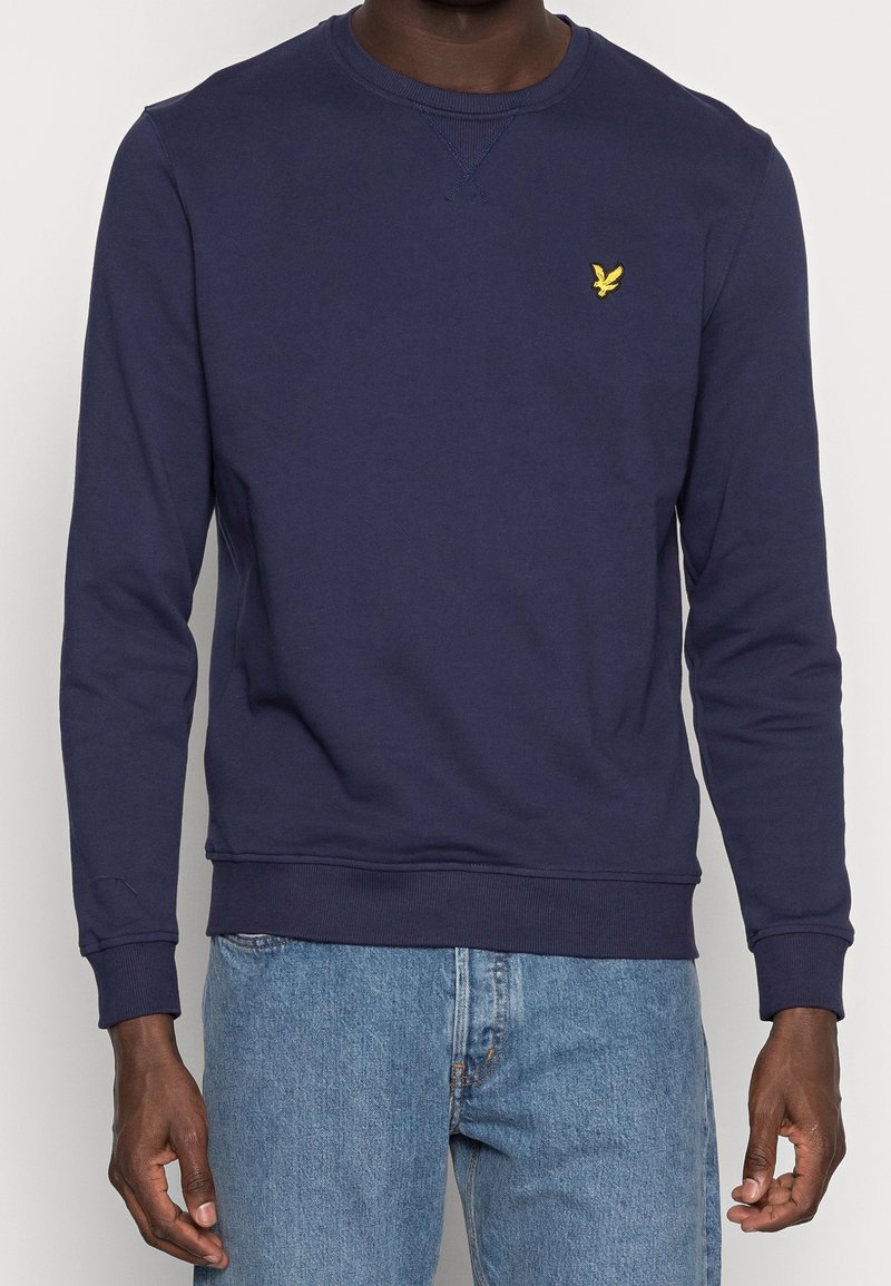 Sudadera azul marino de cuello redondo hecha de tela suave, con puños y dobladillo acanalados. Presenta un pequeño logo amarillo en el pecho.