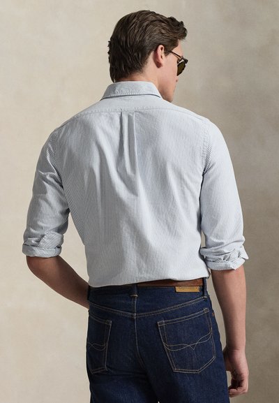 Άνδρας που φοράει ανοιχτό μπλε ριγέ πουκάμισο με σηκωμένα μανίκια και σκούρα μπλε τζιν, βλεπόμενος από πίσω μπροστά σε μονόχρωμο φόντο.