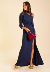 Robe longue navy à une manche, taille cintrée et fente haute. Associée à une pochette rouge et des talons plats dorés. Texture de tissu lisse.