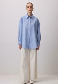 Donna in piedi contro uno sfondo neutro, indossa una camicia oversized a strisce blu e bianche, pantaloni larghi color crema e scarpe bianche.