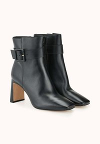 Bottines en cuir noir avec un bout carré, un talon carré, une fermeture éclair et une sangle décorative à boucle autour de la cheville. Texture lisse.
