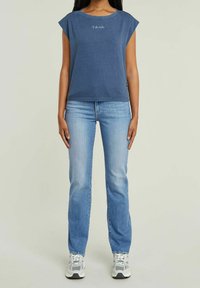 Žena v modré bezrukávové košili, světlých modrých džínách a bílých teniskách, stojící před jednoduchým světlým pozadím.