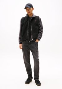 Schwarze Weste über einem grauen Langarmshirt, kombiniert mit dunkelgrauen Jeans und schwarzen Turnschuhen. Die Weste hat einen Reißverschluss und ein Logo.