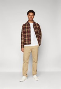 Ruda languota mygtukais užsegama marškiniai su dviem krūtinės kišenėmis, po jais balti marškinėliai, smėlio spalvos kelnės ir balti sportbačiai; visa tai neutralaus fono.