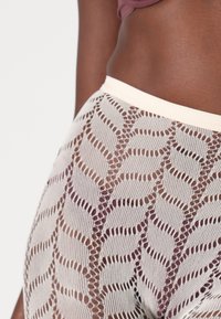 Beige mesh leggings featuring a leaf pattern with open stitches. Elastic waistband and a smooth texture. Close-up view of the waistband and design.