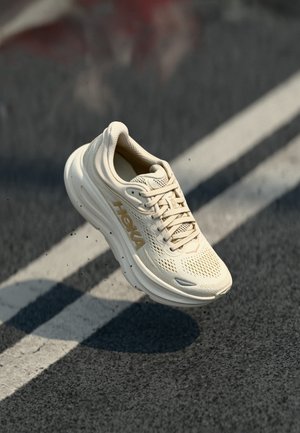Beige Laufschuh mit Mesh-Obermaterial, runder Zehenpartie, strukturiierter Sohle und auffälligem Logo. Leichtes Design mit Schnürsenkeln und Polsterungsdetails.