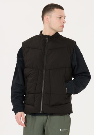 Junger Mann trägt eine schwarze gesteppte Weste über einem schwarzen Langarmshirt, mit grüner Hose, schaut nach rechts vor einem einfarbigen Hintergrund.