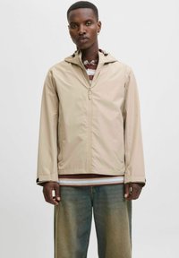 Beige lettvektsjakke med glidelås foran, justerbar hette og elastiske mansjetter. Brukt over en stripete skjorte og kombinert med avslappede jeans.