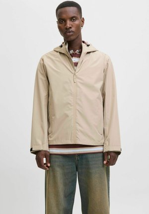 Beige lättviktsjacka med dragkedja fram, justerbar huva och elastiska mudd. Bärs över en randig tröja och kombineras med avslappnade jeans.