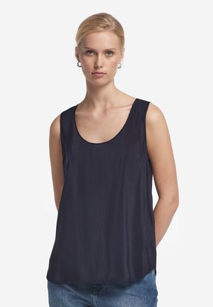 Kvinde iført en sort ærmeløs top med rund halsudskæring og blå jeans, stående mod en ensfarvet hvid baggrund.