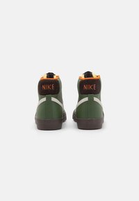 Zapatillas altas verdes con acentos en negro y naranja, material de cuero, parte posterior texturizada y prominente logo "NIKE" en el talón.
