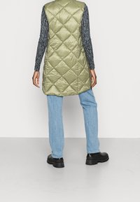 Jasnozielona, pikowana długa kamizelka z wysokim kołnierzem i bocznymi rozcięciami, zestawiona z wzorzystą bluzką z długim rękawem oraz niebieskimi dżinsami. Czarne, masywne buty.