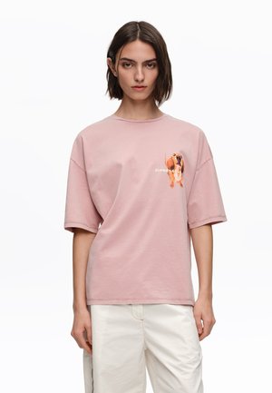 Mujer con cabello castaño corto que lleva una camiseta rosa de gran tamaño con un dibujo pequeño de un perro y pantalones color blanco roto, posando frente a un fondo blanco.