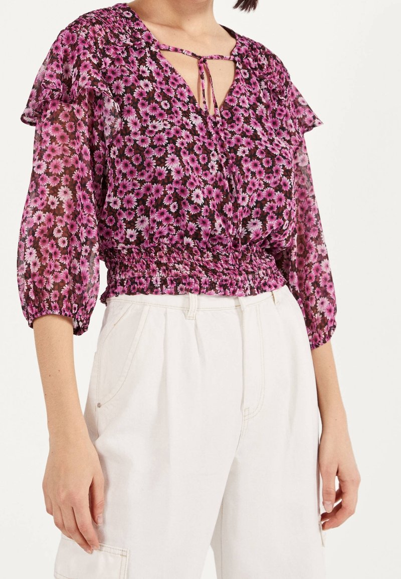 Zbliżenie na osobę ubraną w fioletową bluzkę w kwiaty z rękawami 3/4 oraz białe spłaszczone spodnie na tle jednorodnym.
