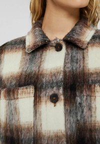 Knappad flanelljacka i kräm, brun och grå rutig mönster. Har krage och bröstficka, tillverkad av texturerat ullmaterial.