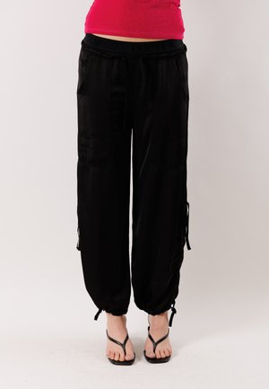 Donna che indossa pantaloni cargo neri a taglio largo con polsini elasticizzati e sandali neri con tacco, in piedi contro uno sfondo semplice.