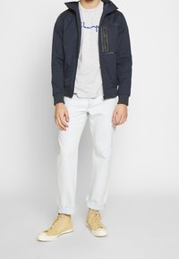 Marinblå zip-up jacka med huva, grå t-shirt med blå text, ljusblå jeans och beige hög-top sneaker. Ren design, avslappnad stil.