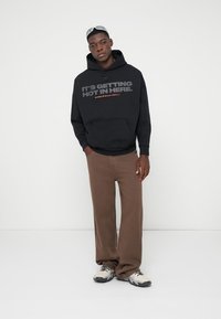 Μαύρη μα hoodie με κείμενο σε γκρι και πορτοκαλί, καφέ παντελόνι φαρδιάς γραμμής, και γκρι αθλητικά. Το μοντέλο στέκεται μπροστά σε λευκό φόντο.