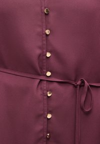 Μπορντό ύφασμα με κάθετη σειρά από μικρά στρογγυλά χρυσά κουμπιά και λεπτή ζώνη στο ίδιο χρώμα, δεμένη κόμπο στη μέση.