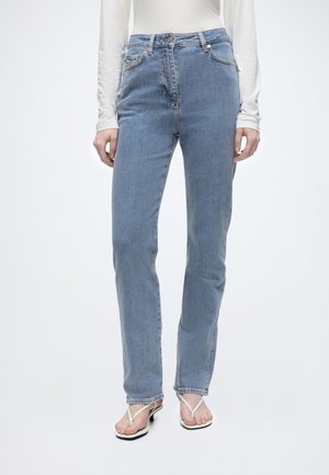 Femme portant un jean droit taille haute bleu clair, un top blanc à manches longues et des sandales blanches à talons, debout devant un fond uni.