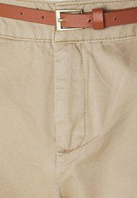 Beige-housut, joissa on sileä kangas, näkyvät ompeleet ja ruskea nahavyö, jossa on kullanvärinen solki vyötäröllä.