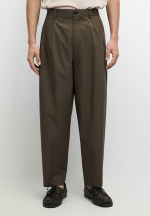 Mann trägt hoch taillierte, locker sitzende braune Hosen mit schwarzen Schnürschuhen, steht vor einem schlichten hellen Hintergrund.